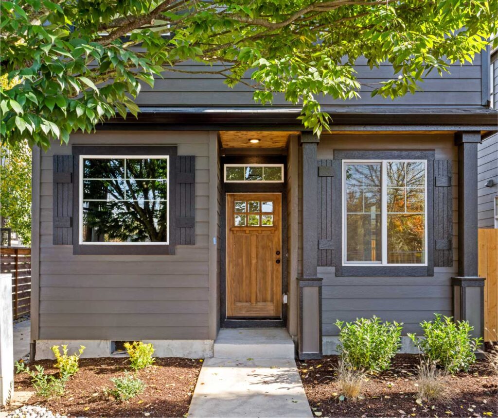 WEB-GreenwoodHomesPDX_AboutCommunity Front entrance of a gray house with a wooden door, two windows with dark shutters, and a small landscaped garden. References a tree canopy above.