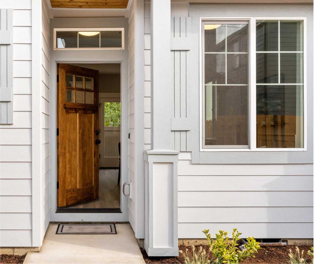 WEB-GreenwoodHomesPDX-_AboutCommunity3 Open wooden front door of a light-gray house, showing a small entry porch and a large window to the right.